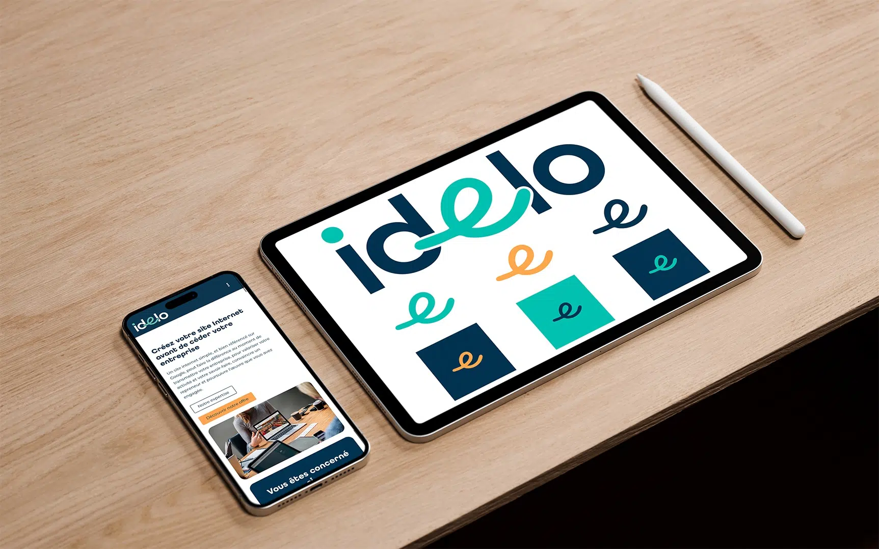 Sur une table , écran de tablette graphique présentant le logo Idelo et ses déclinaisons avec un stylet pour dessiner sur Procreate, accompagné d'un téléphone portable présentant le site internet de Idelo
